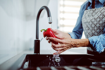 Close up of caucasian senior woman in apron washing red pepper in kitchen sink. - Powered by Adobe