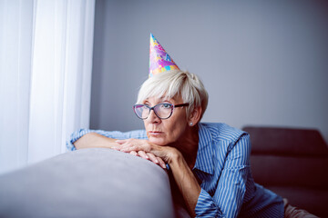Sad beautiful Caucasian senior woman with eyeglasses and birthday cap on head leaning on sofa and...