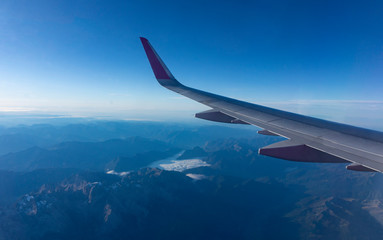 The view from the window of a passenger airliner flying over the territory of the Alps mountain range. Airplane wing on a background of snow-capped mountains in a clear morning sky