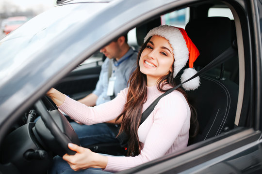 Picture of attractive young woman sit on driver's place. Hold hands on steering wheel and smile. Christmas or new year time. Wear red hat and smile. Man sit beside.
