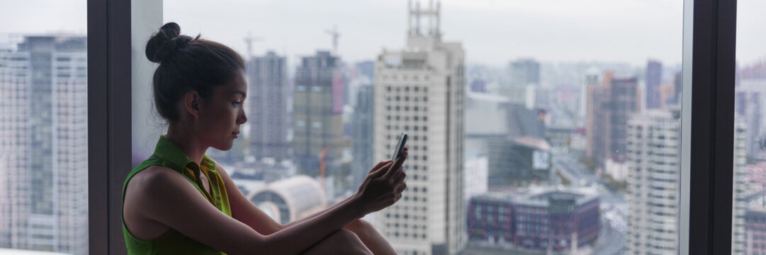 Asian business woman using mobile phone texting on city background at window view of office building or luxury high rise building panoramic banner background.