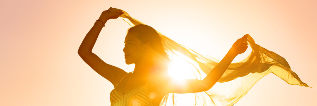 Beautiful woman happy free in sunset glow waving scarf in the wind dancing in sun panoramic banner. Silhouette feminine lady freedom for wellness and healthy living.