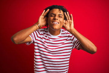 Young afro man with dreadlocks wearing striped t-shirt standing over isolated red background Trying to hear both hands on ear gesture, curious for gossip. Hearing problem, deaf