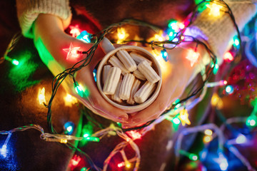 Girls hands hold cup of homemade hot chocolate with white marshmallows, a lot of lights of stars garland and blanket, christmas and new year concept