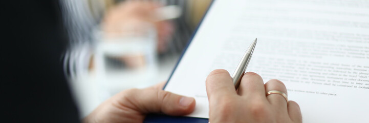 Focus on male hand holding pen and paper folder with contract. Partner checking conditions of important biz bargain in meeting room. Blurred background