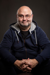 Obraz premium A smiling bald man with glasses sits. Black background. Vertical.