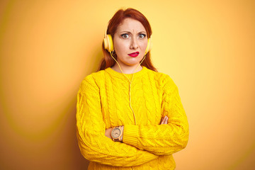 Young redhead woman listening to music using headphones over yellow isolated background skeptic and...