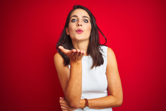 Young beautiful woman wearing white dress standing over red isolated background looking at the camera blowing a kiss with hand on air being lovely and sexy. Love expression.