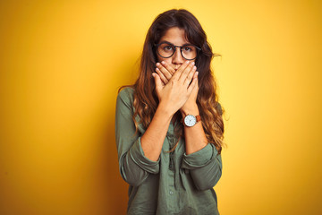 Young beautiful woman wearing green shirt and glasses over yelllow isolated background shocked covering mouth with hands for mistake. Secret concept.