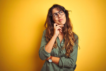 Young beautiful woman wearing green shirt and glasses over yelllow isolated background with hand on chin thinking about question, pensive expression. Smiling with thoughtful face. Doubt concept.