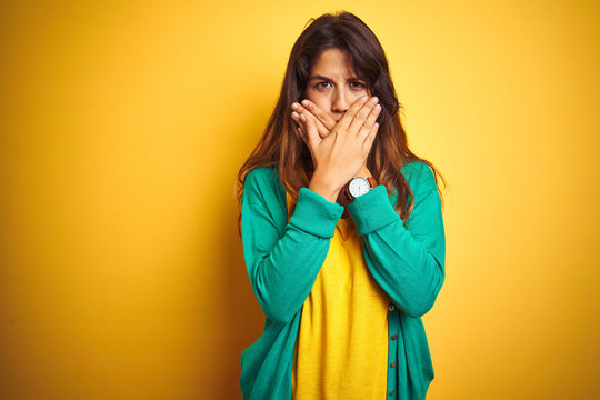 Young woman wearing t-shirt and green sweater standing over yelllow isolated background shocked covering mouth with hands for mistake. Secret concept.
