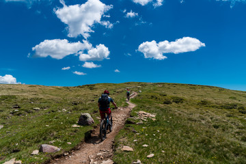 Fototapeta premium Zwei Mountainbiker auf einem Trail zwischen Wiesen aufwärts mit blauem Himmel und weißen Wolken