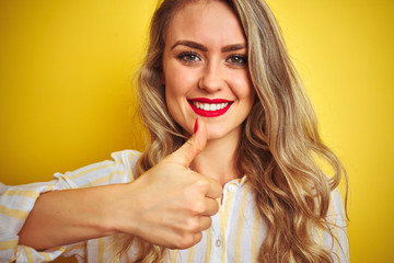 Young beautiful woman wearing stripes shirt standing over yellow isolated background happy with big smile doing ok sign, thumb up with fingers, excellent sign