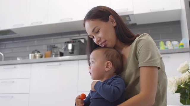 Low angle medium shot of young Asian woman sitting on floor in kitchen at home, hugging and kissing baby, talking and looking at camera with happy face