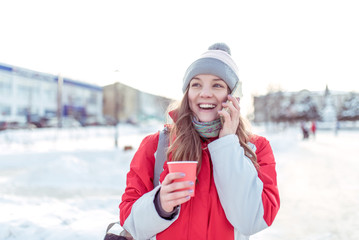 Happy laughs, smiles, I rejoice. Beautiful girl in winter on street makes phone call, her hand cup of coffee tea, breakfast snack on nature. Warm jacket hat. Listens message online on Internet.