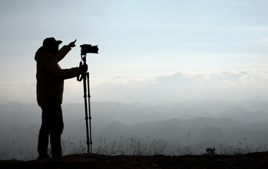 Silhouette image of photographer hold camera with tripod and point to the mountain over the cliff with morning light.