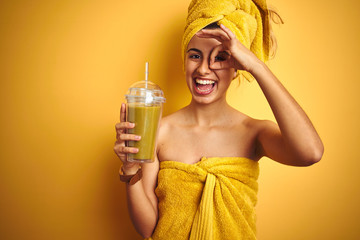 Young beautiful woman wearing a towel drinking detox juice over yellow isolated background with happy face smiling doing ok sign with hand on eye looking through fingers
