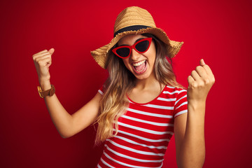 Young beautiful woman wearing sunglasses and summer hat over red isolated background very happy and excited doing winner gesture with arms raised, smiling and screaming for success