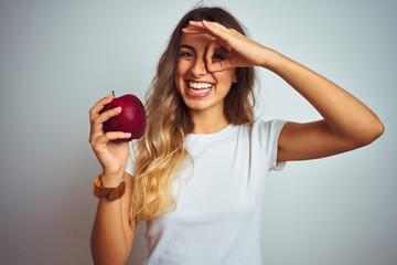 Young beautiful woman eating red apple over grey isolated background with happy face smiling doing ok sign with hand on eye looking through fingers