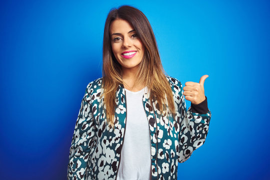 Young beautiful woman wearing a jacket standing over blue isolated background smiling with happy face looking and pointing to the side with thumb up.