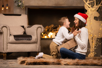 Photo of woman in Santa's cap with son sitting on floor on background of fireplace