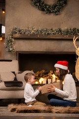 Picture of brunette woman in Santa's cap with son sitting on floor on background of fireplace