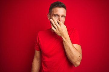 Young handsome man wearing casual t-shirt over red isolated background smelling something stinky and disgusting, intolerable smell, holding breath with fingers on nose. Bad smells concept.