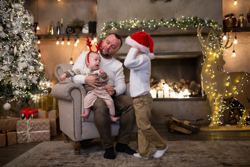 Picture of father with his sons in santa cap sitting in armchair by New Year tree and fireplace
