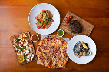 Dining wooden table with food, top view. Pizza, salad, cheese board, steak, black seafood pasta. Concept for restaurant menu.