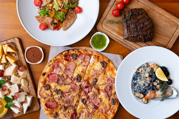 Dining wooden table with food, top view. Pizza, salad, cheese board, steak, black seafood pasta. Concept for restaurant menu.