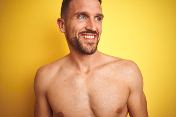 Sexy young shirtless man showing nude fitness chest over yellow isolated background looking away to side with smile on face, natural expression. Laughing confident.