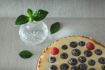 Homemade berry cake with raspberries and blueberries and crystal vase with small bud of gardenia on gray minimalistic background