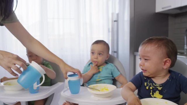 Tracking shot of young Asian mom bringing and giving three sippy cups with water for messy toddler triplets sitting in high chairs in kitchen after lunch