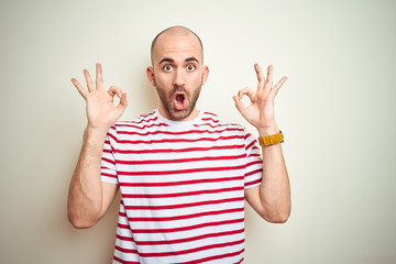 Young bald man with beard wearing casual striped red t-shirt over white isolated background looking surprised and shocked doing ok approval symbol with fingers. Crazy expression