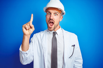 Young handsome engineer man wearing safety helmet over blue isolated background pointing finger up with successful idea. Exited and happy. Number one.