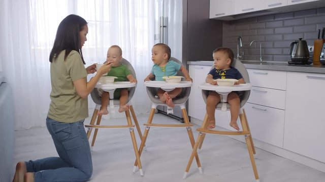 Dollying shot of young Asian mother kneeling in kitchen next to high chairs at home and feeding baby puree to messy 1-year-old toddler triplets with spoon