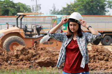 Obraz premium Female civil engineer standing and catch the white helmet on loader pedal car and pile of soil background.