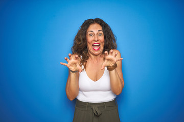 Fototapeta premium Middle age senior woman with curly hair standing over blue isolated background smiling funny doing claw gesture as cat, aggressive and sexy expression