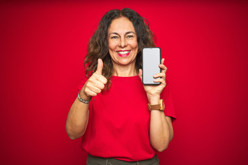 Naklejka premium Middle age senior woman showing screen of smartphone over red isolated background happy with big smile doing ok sign, thumb up with fingers, excellent sign