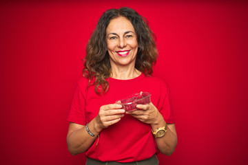 Middle age senior woman eating raspberries over red isolated background with a happy face standing and smiling with a confident smile showing teeth