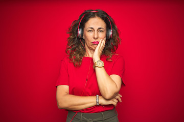 Middle age senior woman wearing headphones listening to music over red isolated background thinking looking tired and bored with depression problems with crossed arms.