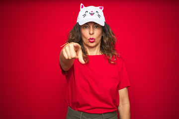 Middle age senior woman wearing cute kitty cap over red isolated background pointing with finger to the camera and to you, hand sign, positive and confident gesture from the front