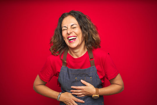 Middle age senior woman wearing apron uniform over red isolated background smiling and laughing hard out loud because funny crazy joke with hands on body.