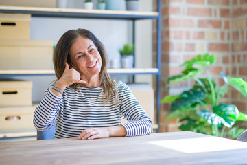 Middle age senior woman sitting at the table at home smiling doing phone gesture with hand and fingers like talking on the telephone. Communicating concepts.