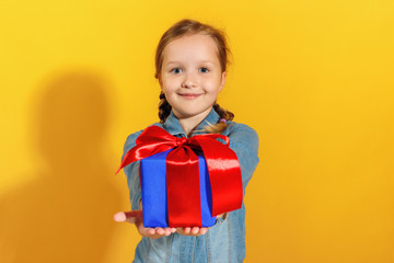 Happy child gives a present. Cute little girl in a denim shirt on a yellow background