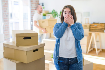 Young couple arround cardboard boxes moving to a new house, plus size woman standing at home shocked covering mouth with hands for mistake. Secret concept.