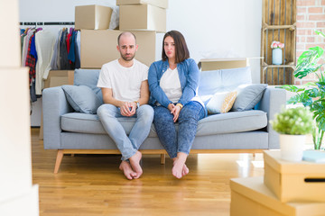 Young couple sitting on the sofa arround cardboard boxes moving to a new house depressed and worry for distress, crying angry and afraid. Sad expression.
