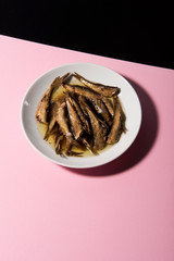 Pile of oily fish on white plate on pink tabletop with black background. Floating fork above. Abstract conception.
