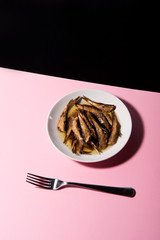 Pile of oily fish on white plate on pink tabletop with black background. Floating fork above. Abstract conception.