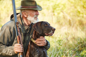 Fototapeta Caucasian mature man with gun and dog sit searching prey. Bearded man in hunting clothes. Autumn obraz